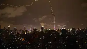 A cityscape with block-like, shadowed towers under a night sky with a bronze glow from a distant illuminated skyline in the background and lightning striking