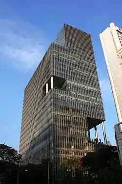 Ground-level view of a 20-storey skyscraper with a brown façade; the building has a rectangular cross section and is cut-off roughly 10 stories up, with the uppermost 10 floors partially protruding out over the street below.