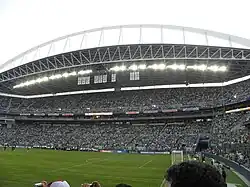 The grandstand of a stadium filled with people. The stadium has distinctive trusses for support.