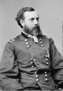 Head and torso of a white man with a full beard, wearing a partially unbuttoned double-breasted military jacket, a handkerchief tied around his neck, and a single medal pinned to his left breast.