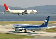 A white and red-tailed Japan Airlines aircraft above the runway, with landing gears down, and an All Nippon Airways in blue and white livery taxiing