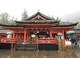 Frontal view of a wooden building with vermillion red beams. A platform with red handrail is located in front of the building.