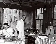 Two scientists in white lab coats, one seated and one standing, in a small building with peeling paint, standing next to a table with scientific glassware