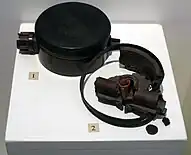 Black disc-shaped landmine on a white display stand, alongside broken fragments of another landmine.