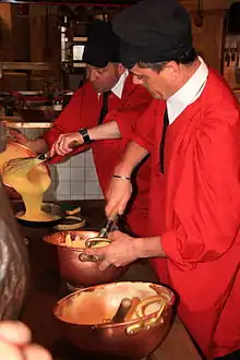 Two chefs in red smocks and black berets, one stirring eggs in a copper bowl and the other pouring batter from a bowl into a pan