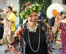 Mary Ama is smiling into the camera with her arms out. She hold a fan in one hand, has a beautiful woven head piece and is looking joyful.