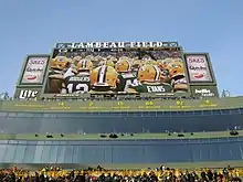 A photo of the stands at Lambeau Field, with a row of the Packers' retired numbers and player names located just above the suite seats. A large video screen is located above the numbers.