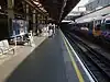 A platform with several people waiting for a train. A white square sign has a London Underground roundel with "PADDINGTON" in the centre. To the right there is a track with fourth track electrification and on an adjacent track a blue multiple unit train with red doors waits.