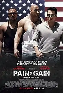 Three musclebound men stand in front of a large American Flag