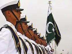 Pakistan Navy soldiers in a straight line are standing next to their national flag.