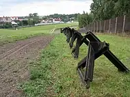 View along a row of triangular metal spikes standing between a bare earth strip and a metal fence.