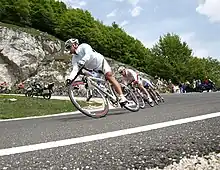 A group of cyclists, in a single-file line, on a road that is descending in elevation. The cyclist in the front of the group wears a white rain jacket, and his shorts bear rainbow stripes.