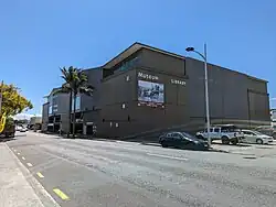 A square brown building has a sign stating 'Museum' and 'Library' and has a large black and white historical photograph under the signs. There is a carpark entrance visible further down the road along the building.