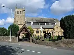 A stone church seen from the south. The tower on the left has a clock face, a flagpole and a weather vane; the body of the church has a timber-framed clerestory. In front of the church is a churchyard with gravestones, a crucifix, a lych gate, a notice board, a yew tree, and a wall. A road runs through the foreground with a lamppost to the left of the church tower