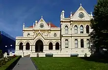 A sunlit grand building is upon a sloping green lawn. It has a big staricase leading teh the entrance, several floors and is made of stone in an old fashioned English sort of style. There are flags on flagpoles.