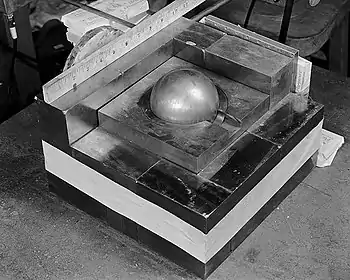 A stack of square metal plates with a side about 10 inches. In the 3-inch hole in the top plate there is a gray metal ball simulating Pu.