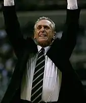 A man raising his hands in the air during a basketball game. He is wearing an open jacket with a white shirt and striped tie.