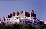 Fortress of dark stones on a hill above a town with white houses.