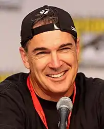 A closeup of a man with his head looking forward, smiling and wearing a backwards baseball cap.