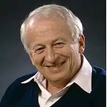 Late middle-aged man with grey hair, wearing a navy blue sweater and a white dress shirt