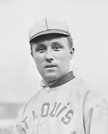 A photo of a man standing up wearing his baseball uniform. He has a baseball in his right hand, and his catcher's mitt in his left. He is in the middle of the field, while two other ballplayers are talking in the background. His facial expression indicates that he did not expect to be photographed.