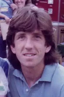 A man sporting a popular 1970s hairstyle, wearing a blue shirt.