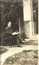 Photograph of Paul Milliet, older than during the events of the Commune. He is sitting on a chair outside a house, holding a book in his hands.