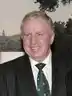 Head and shoulders of Paul Murphy, stood in a dark suit with a shirt and a green tie, in front of a white wall.