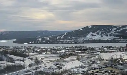 An aerial view of a small settlement beside a wide river, covered in a layer of snow. A large forested hill lies on the other side of the river.