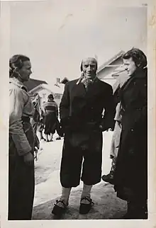 Black and white image of Per Samuelshaug at approximately 33 years old. He is stood stood in the snow with two women. He is wearing a black coat and black trousers with a white hat and white socks