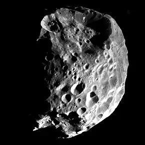 An approximately spherical heavily cratered body is illuminated from the bottom-right. The terminator runs near the left and top limbs. There is huge crater at the top, which affects the shape, and another slightly smaller at the bottom.