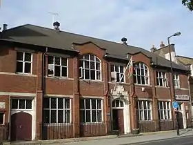 Two storey red-brick building, with seven sets of windows on the upper floor, and for sets on the ground floor. It has three entrance doors, all shut. Railings separate the building from the pavement. A Welsh flag flies from above the central main entrance.