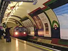 A red train running on a railway track through the interior of a building that has a rounded ceiling and rounded walls covered in advertisements