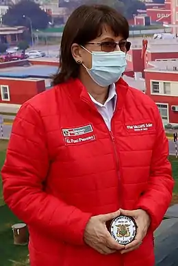 Pilar Elena Mazzetti Soler wearing a red puffer jacket as well as a blue surgical mask and tinted glasses, she is holding a badge