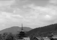 Shot of a Japanese pagoda (in Kyoto), set among ordinary rooftops and trees, with tree-lined mountains and sky in the background.