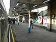 A railway platform with various people walking in different directions on it and a black sign reading "Way out" in yellow letters