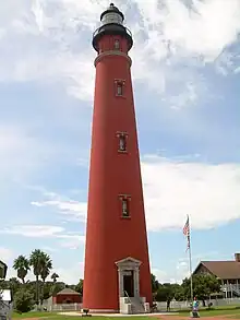 A tall red lighthouse, seen from the point of view of someone on the ground, is seen framed by a blue sky, with palm trees and a flagstaff in the background.