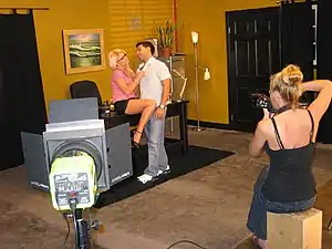 A blonde haired woman wearing a pink pantyand black miniskirt is seated atop an office desk. She seductively tugs on a man's shirt, who is standing between her thighs. In the foreground a second woman sits and photographs them, beside her is a stage light pointed towards the couple.