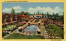 A beautiful garden, green and with white, red, pink, and orange flowers. A reflecting pool is in the center of this photo flanked with four black flowerpots with white flowers at each corner. In the background is a white building with a brown roof.