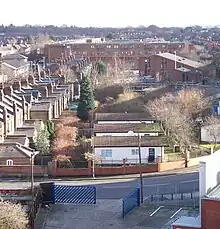 A strip of grass recedes in a straight line into the distance towards a large yellow brick building. Three single-storey huts stand on the grass.