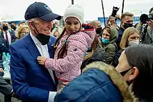 Photo of a smiling Biden holding a child, with a mask lowered onto his chin