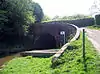 The canal enters the tunnel through and arch and on the right a road curves towards the left to cross the bridge.