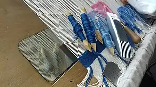 Tapestry tools, on the loom. Bobbins, scrapper (with short teeth), comb (double-ended), and awl (tip hidden).