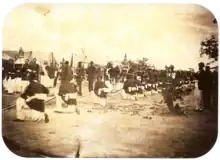 An old photograph showing a procession passing between lines of soldiers with tents in the background