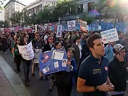 People peacefully marching up a street. Several are carrying signs displaying pro-same-sex marriage slogans, such as "We all deserve the freedom to marry." Most people have sad or serious facial expressions.