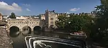 A three arch stone bridge with buildings on it, over water. Below the bridge is a three step weir and pleasure boat.