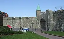 Quebec's restored city wall is gray stone about 20 feet (6.1 meters) high. The St. John's gate has a modern road going through it, and has a copper-roofed turret on the left bastion. A paved path goes through a grassy area below the wall.