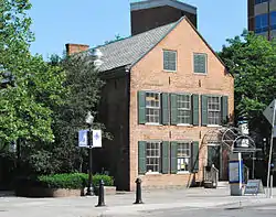 Side view of two-and-a-half-story brick house with wooden shutters next to the windows in an urban setting. There are trees next to it on the left.