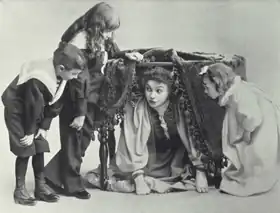 young women peering out from under a table, through the folds of the tablecloth; she is surrounded by three small children