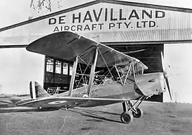 Biplane on tarmac in front of hangar marked "De Havilland Aircraft Pty Ltd"